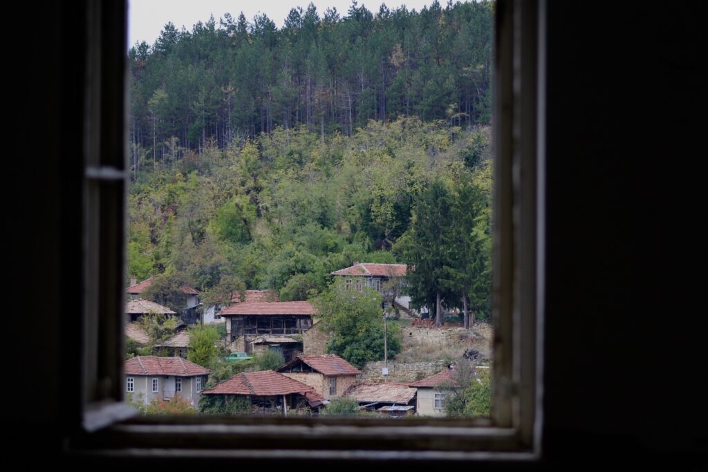 An image of a window view which shows a hill with trees and houses in a Bulgarian village.